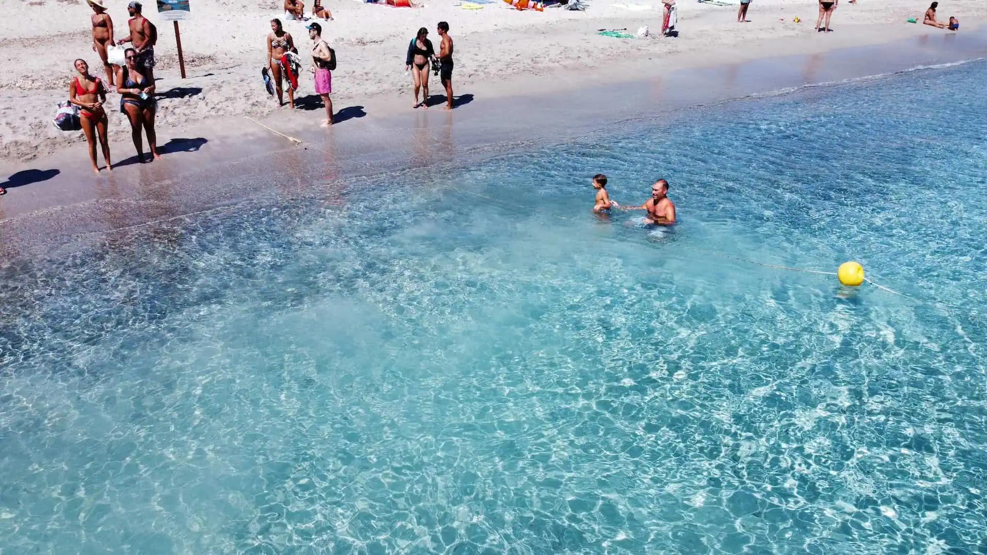 A father and child enjoying the shallow, crystal-clear waters near a white sandy beach, surrounded by beachgoers.