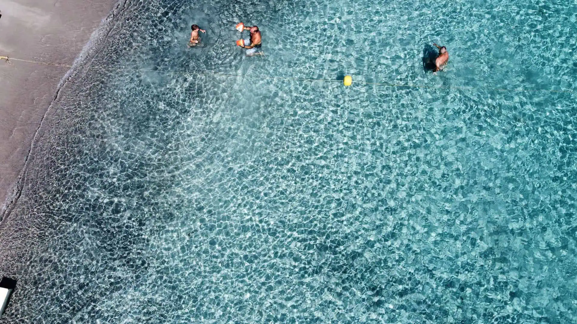 Aerial view of people swimming in crystal-clear shallow water near the shore, with sunlight creating rippling patterns on the seabed.