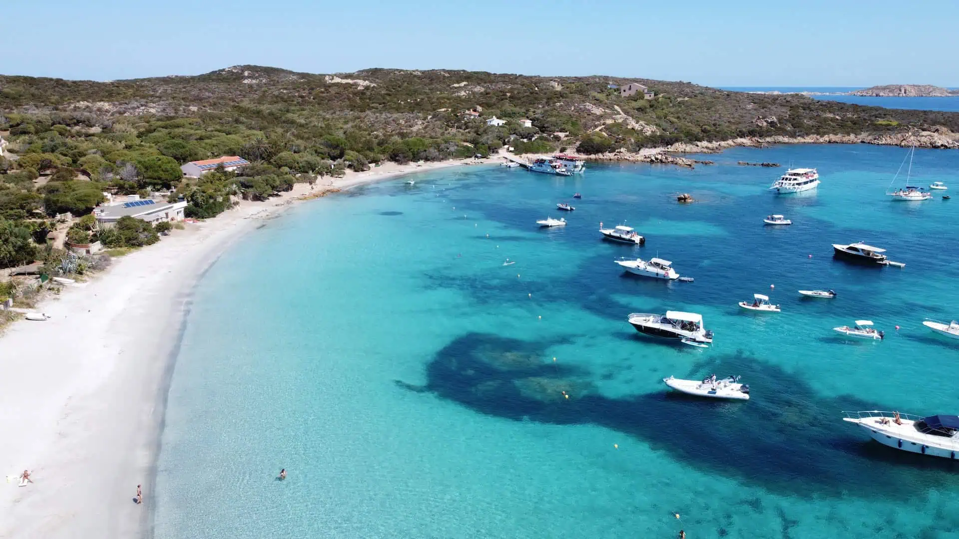A stunning aerial view of a turquoise bay with white sandy beaches, lush greenery, and numerous boats anchored in the clear waters.