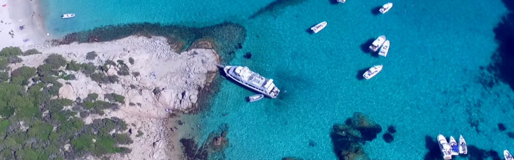 An aerial view of a rocky coastline with a yacht anchored in shallow turquoise waters, surrounded by smaller boats.