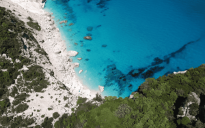 Aerial view of a secluded beach in North Sardinia, featuring turquoise water, a rocky shoreline, and lush green vegetation on the cliffs.